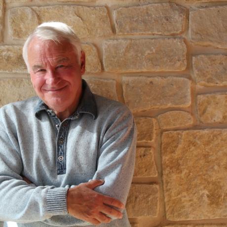 Tom Golisano, a smiling man wearing a gray-collared sweater stands against a stone wall with his arms crossed.